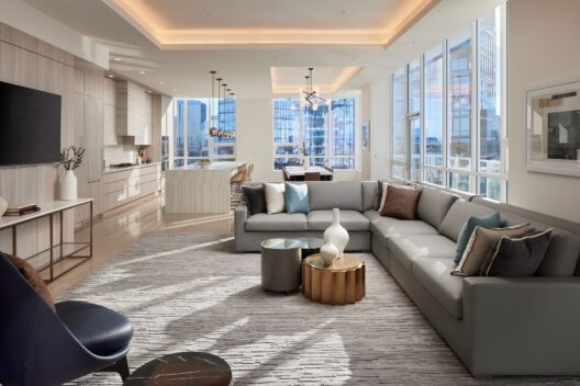 A modern, sunlit living room with floor-to-ceiling windows, a large gray sectional sofa, stylish accent tables, and open kitchen area, featuring neutral tones, contemporary decor, and cityscape views outside.