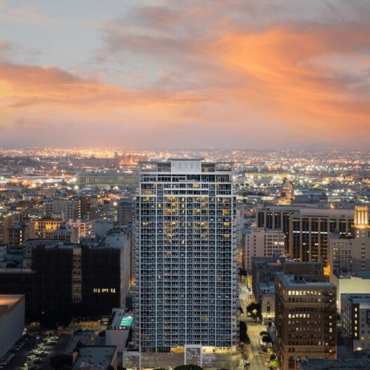 A tall, modern high-rise building stands illuminated in the center of a cityscape at sunset, with a dramatic orange sky and city lights stretching into the distance.