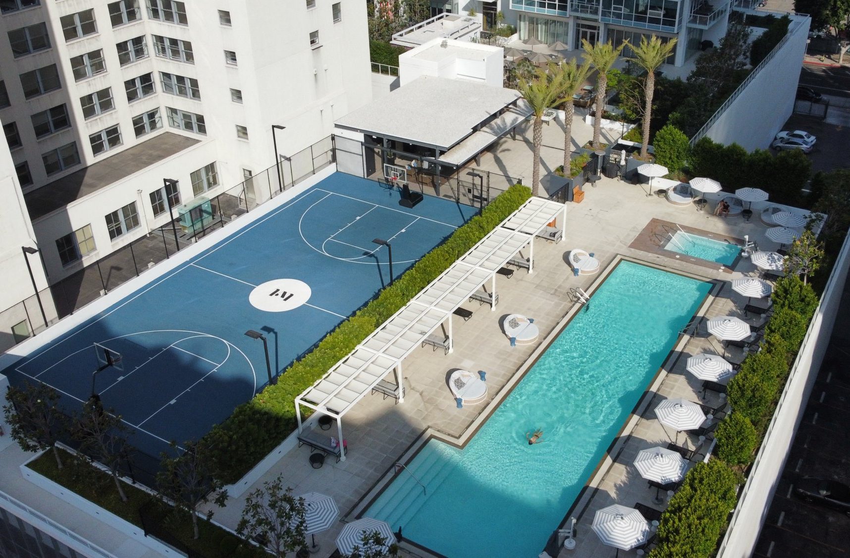 A rooftop view of an outdoor area with a blue basketball court, a rectangular swimming pool, sun loungers, white umbrellas, palm trees, and surrounding modern buildings.