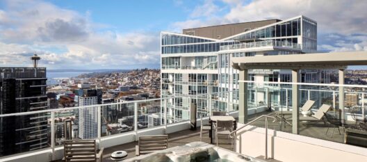 Rooftop patio with lounge chairs, tables, and a hot tub, surrounded by glass railings overlooking a cityscape with modern buildings, hills, and the Space Needle in the distance under a partly cloudy sky.