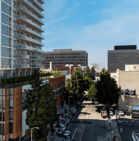 A city street lined with trees, parked cars, and modern office buildings under a clear blue sky. A tall glass building is on the left, with mid-rise buildings and greenery visible throughout the scene.