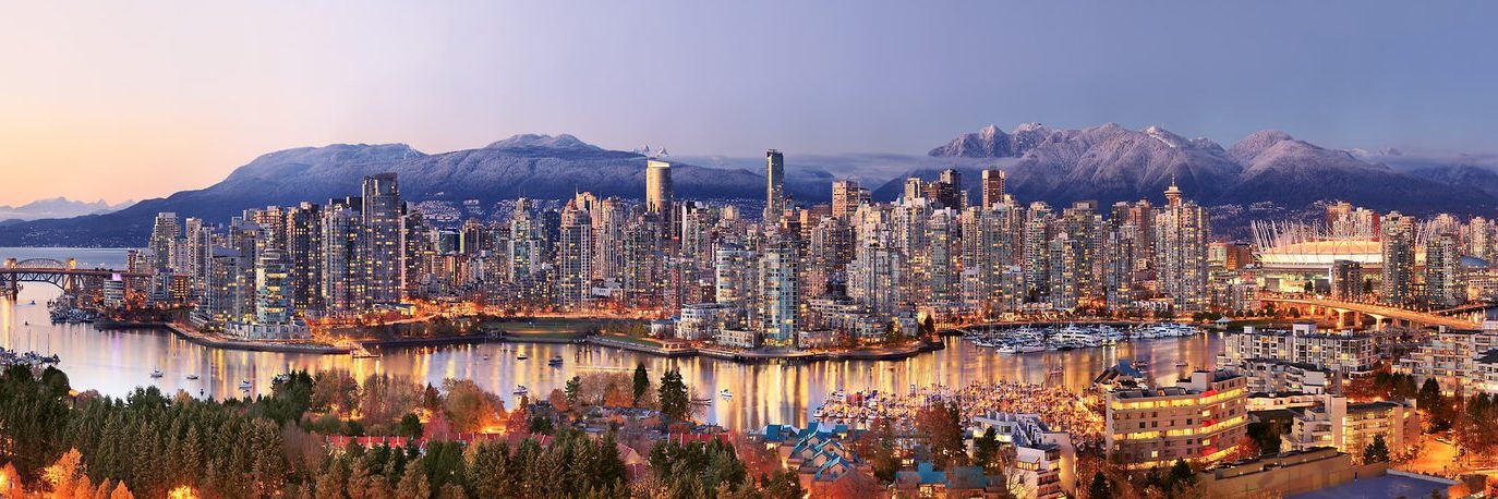 A panoramic view of Vancouver’s skyline at dusk, with modern skyscrapers, illuminated buildings, and snow-capped mountains in the background reflecting in the calm harbor waters.