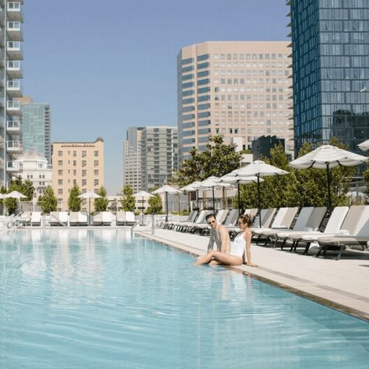 Two people sit by the edge of a clear outdoor pool surrounded by lounge chairs and umbrellas, with tall modern buildings and a blue sky in the background.
