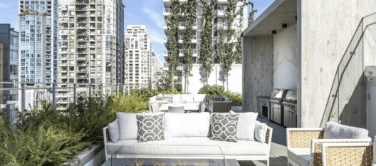 A modern rooftop patio with white sofas, patterned pillows, wicker chairs, a glass railing, greenery, and built-in grills. Tall city buildings surround the space under a clear blue sky.