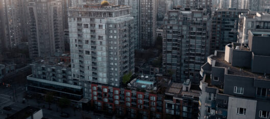 Aerial view of tall apartment buildings in a city, with a rooftop swimming pool visible in the center among the gray high-rises. Streets and parked cars are seen below, and the urban area appears densely populated.