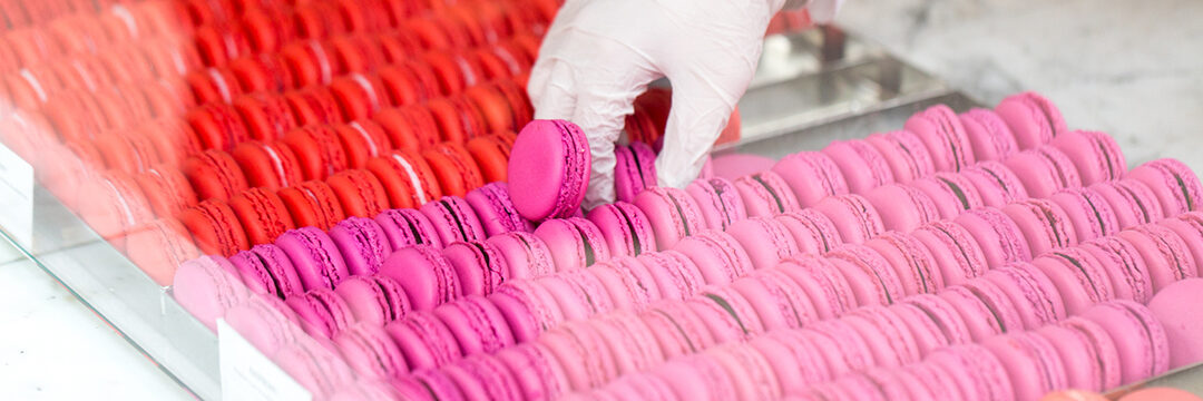 A gloved hand selects a pink macaron from a neatly arranged row of pink and red macarons displayed in a glass case at a bakery.