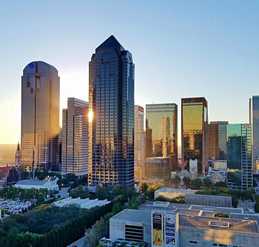 A city skyline at sunset featuring tall glass skyscrapers reflecting sunlight, with smaller buildings, green trees, and clear blue sky in the background.