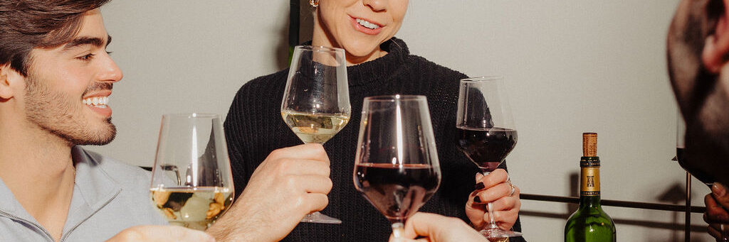 Three people smiling and clinking wine glasses together in a toast, holding both red and white wine. A bottle of wine is visible in the background on a table.