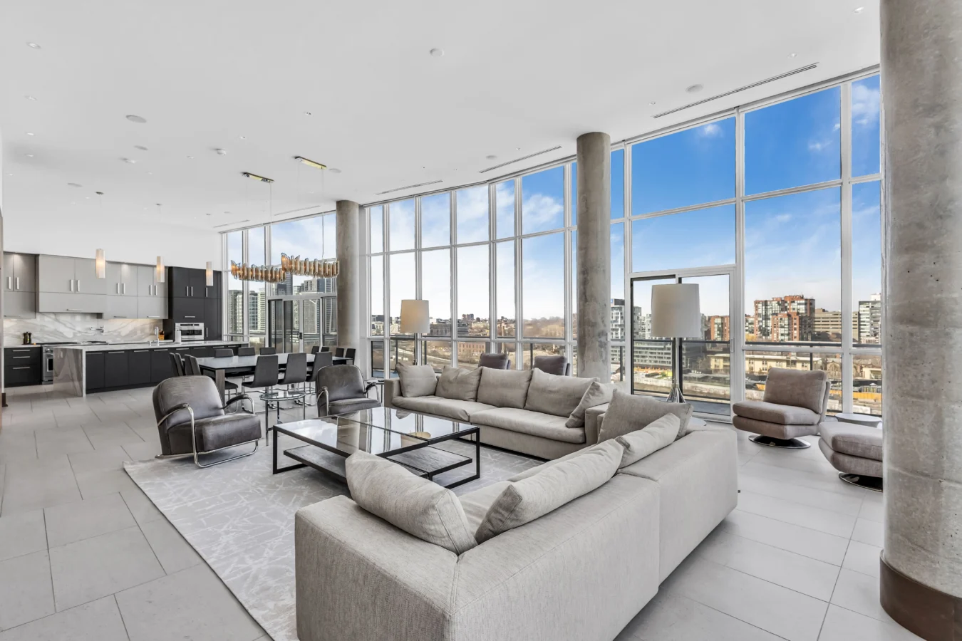 Spacious modern living room with large beige sectional sofa, glass coffee table, and floor-to-ceiling windows offering city skyline views. Open-concept layout includes a sleek kitchen and dining area with contemporary furnishings.