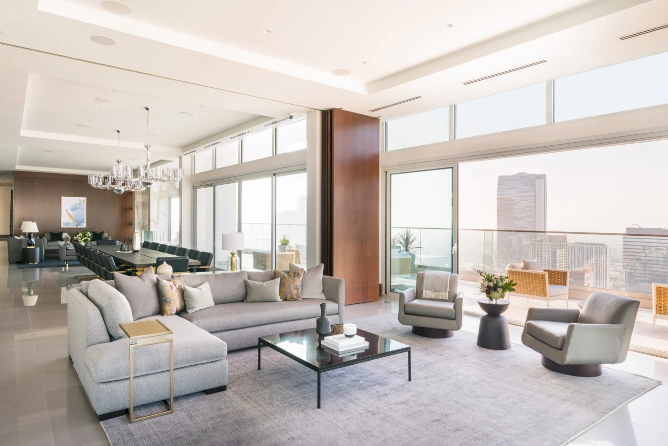 A modern, spacious living room with large windows, neutral-toned furniture, a grey sectional sofa, armchairs, coffee table, and city views. Adjacent dining area with a long table and chandelier. Balcony visible outside.
