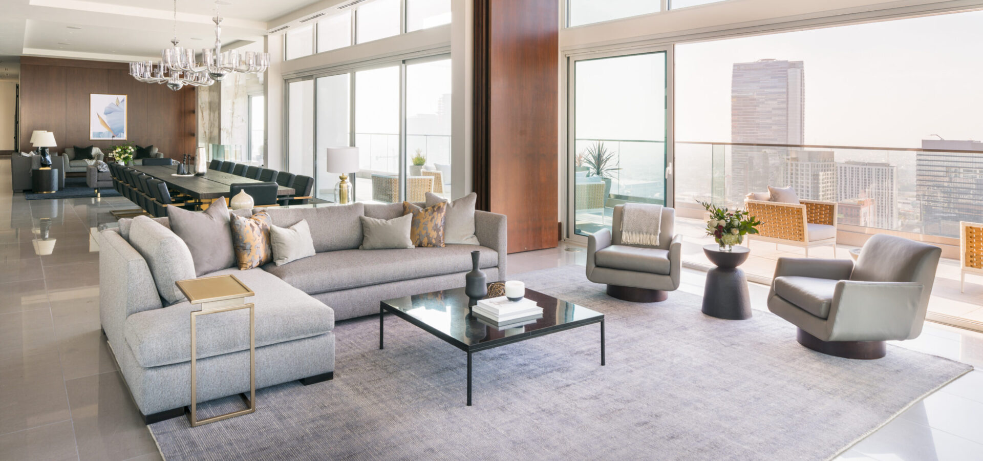 A modern, spacious living room with large windows, neutral-toned furniture, a grey sectional sofa, armchairs, coffee table, and city views. Adjacent dining area with a long table and chandelier. Balcony visible outside.