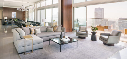 A modern, spacious living room with large windows, neutral-toned furniture, a grey sectional sofa, armchairs, coffee table, and city views. Adjacent dining area with a long table and chandelier. Balcony visible outside.