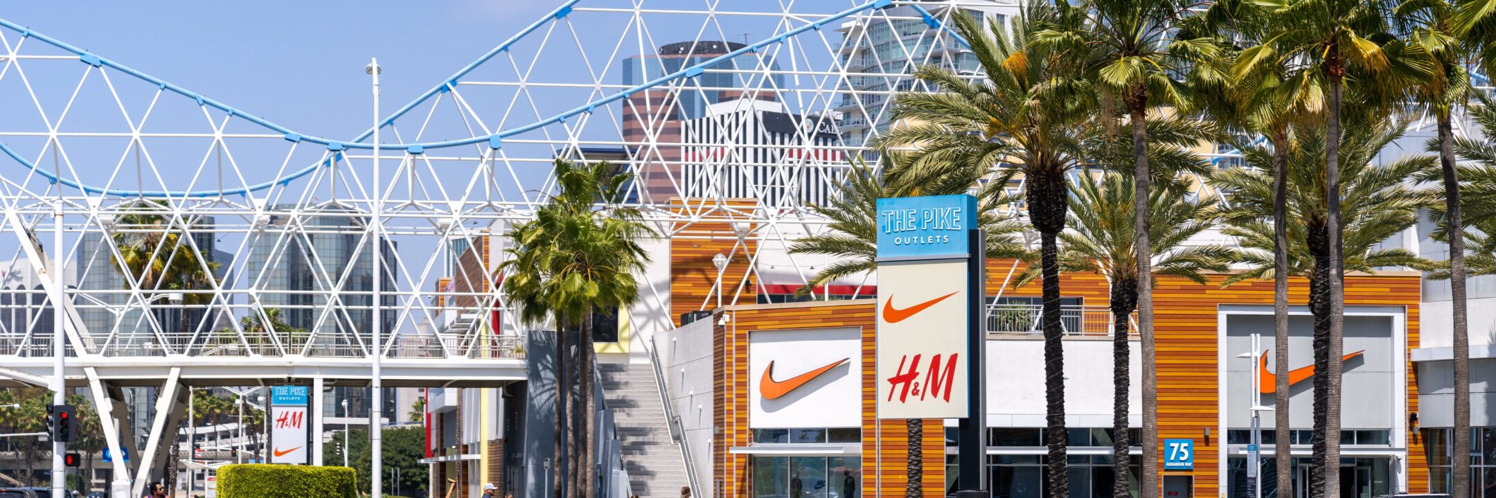 Outdoor shopping area with palm trees, a Nike and H&M store, signs for The Pike Outlets, and a modern white geometric pedestrian bridge under a blue sky.