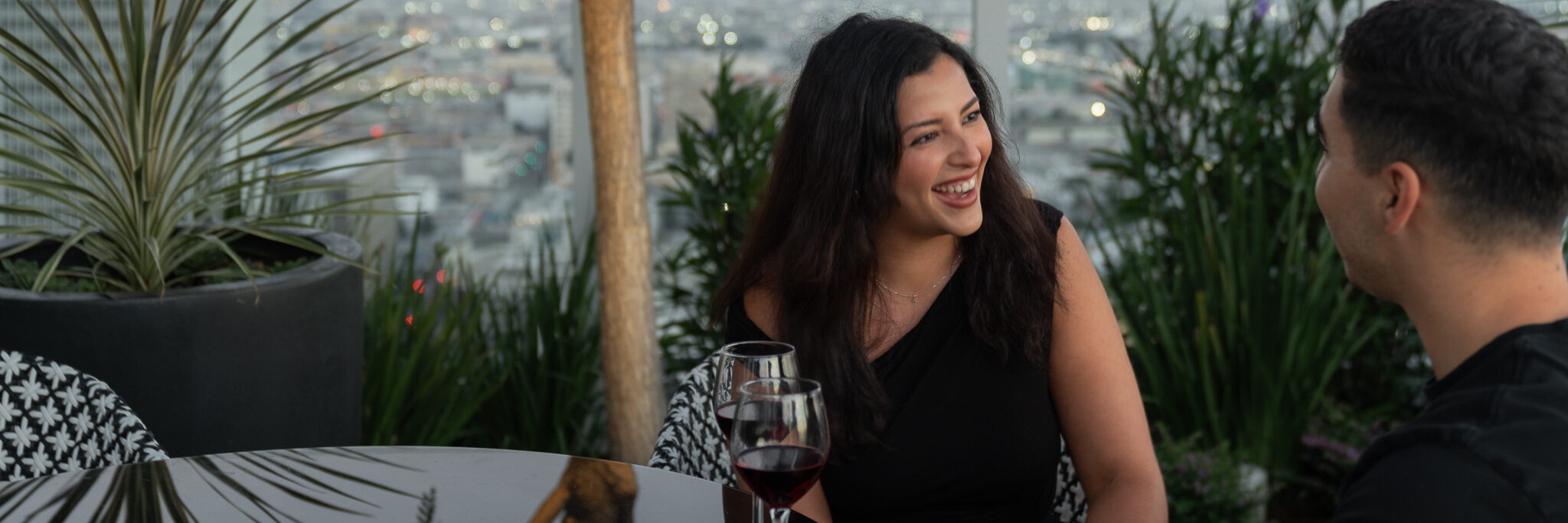 A woman and a man sit at an outdoor table with glasses of red wine, smiling and talking, surrounded by green plants with a blurred cityscape in the background.