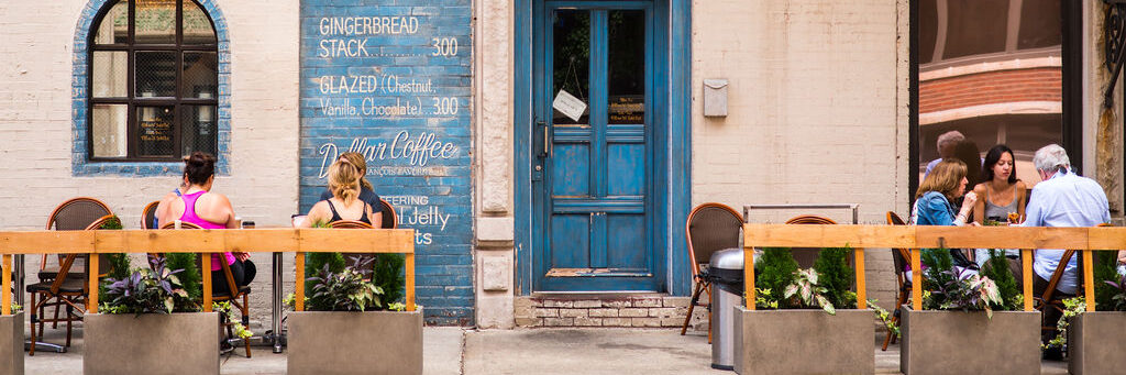 People sit at outdoor tables in front of a café with a blue door and menu painted on the wall. The seating area is lined with planters, and the atmosphere is casual and relaxed on a sunny day.