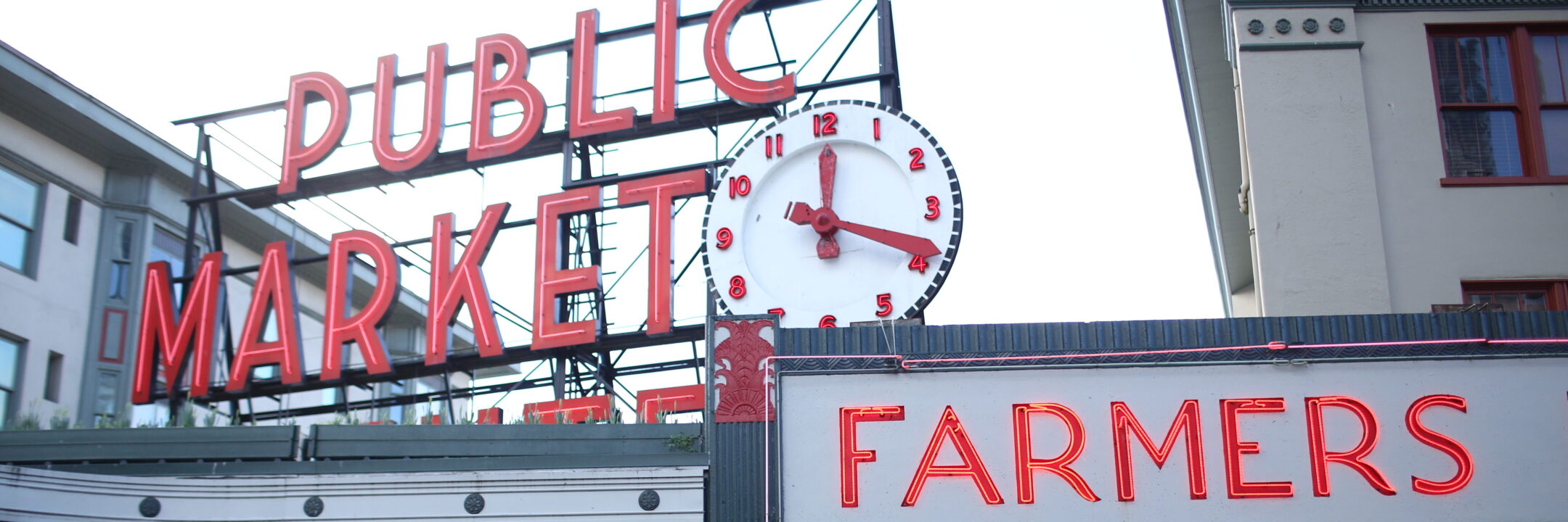 Large neon red Public Market sign and a clock showing 2:34 above the market, with a Farmers sign below. Buildings surround the signs, likely at Pike Place Market in Seattle.