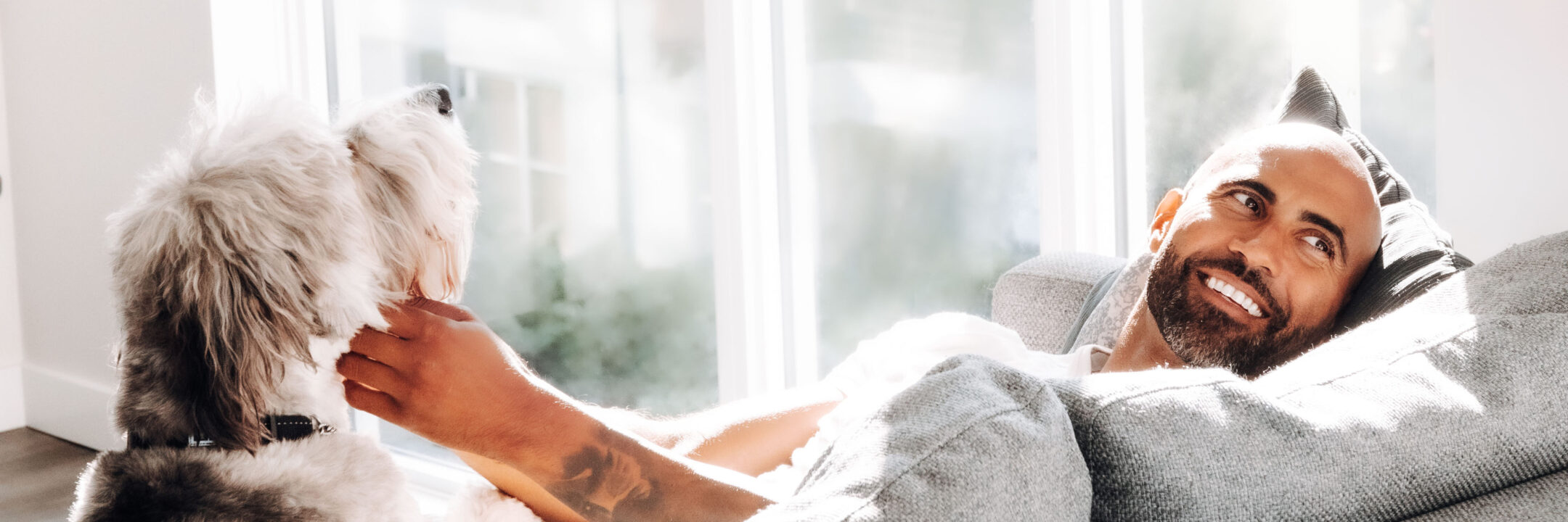 A man relaxes on a couch by a bright window, smiling while petting a fluffy dog sitting beside him.