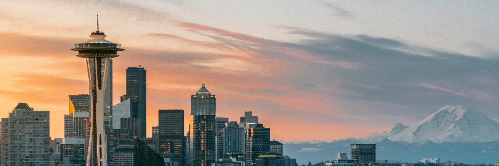 Seattle skyline at sunset with the Space Needle on the left, skyscrapers in the center, and Mount Rainier visible in the background under a colorful sky.