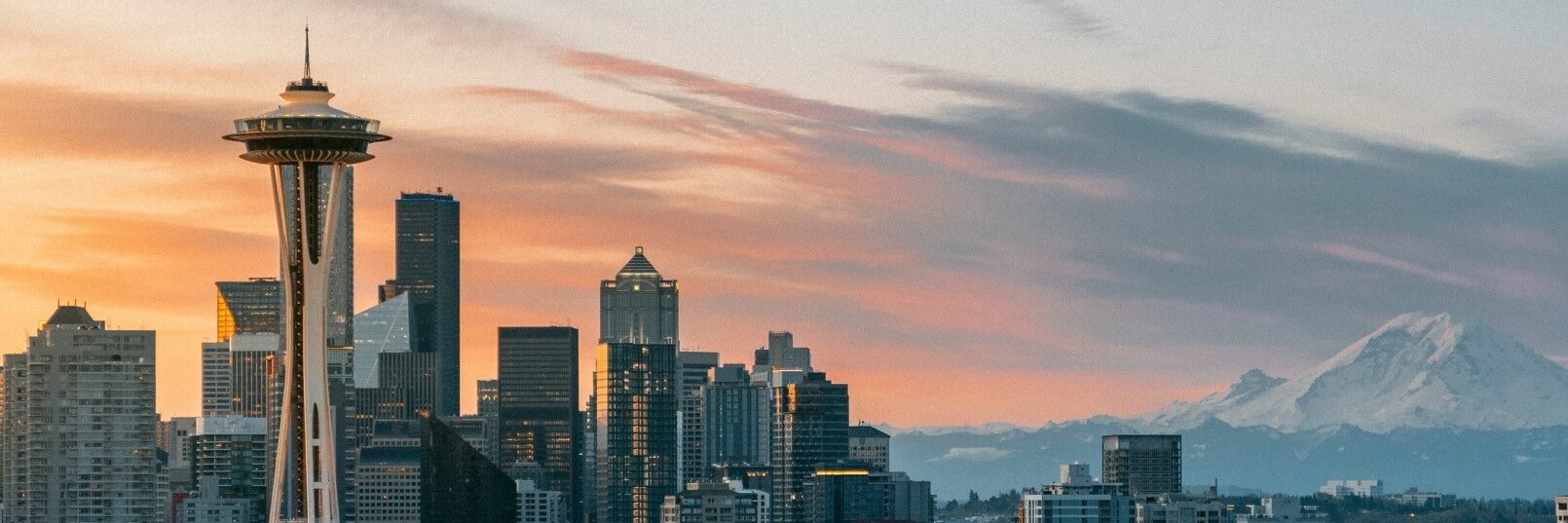 Seattle skyline at sunset with the Space Needle on the left, modern skyscrapers in the center, and snow-capped Mount Rainier in the background under a colorful sky.