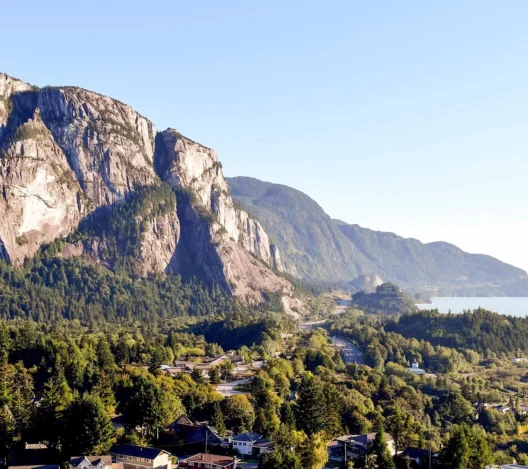 A scenic view of a rocky mountain with forested slopes, a small town in the foreground, and a body of water in the distance under a clear blue sky.