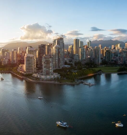 Aerial view of a modern city skyline with tall buildings by the waterfront, boats on the water, green spaces, and mountains visible in the background under a partly cloudy sky.